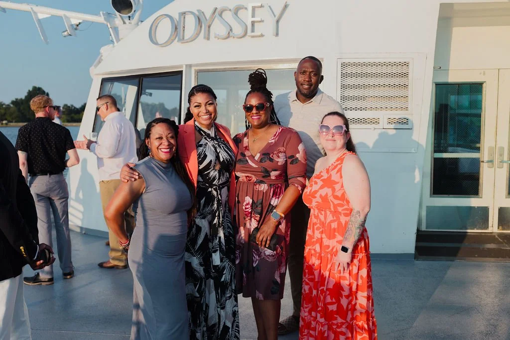 Group of five diverse people smiling and posing in front of a white boat named 'Odyssey', on a sunny day.