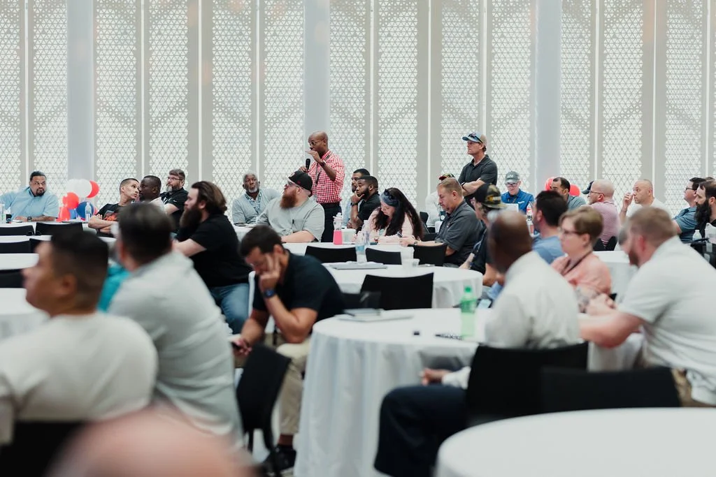 A diverse group of people attending a conference or seminar in a modern, well-lit room, with some taking notes and one person speaking into a microphone.