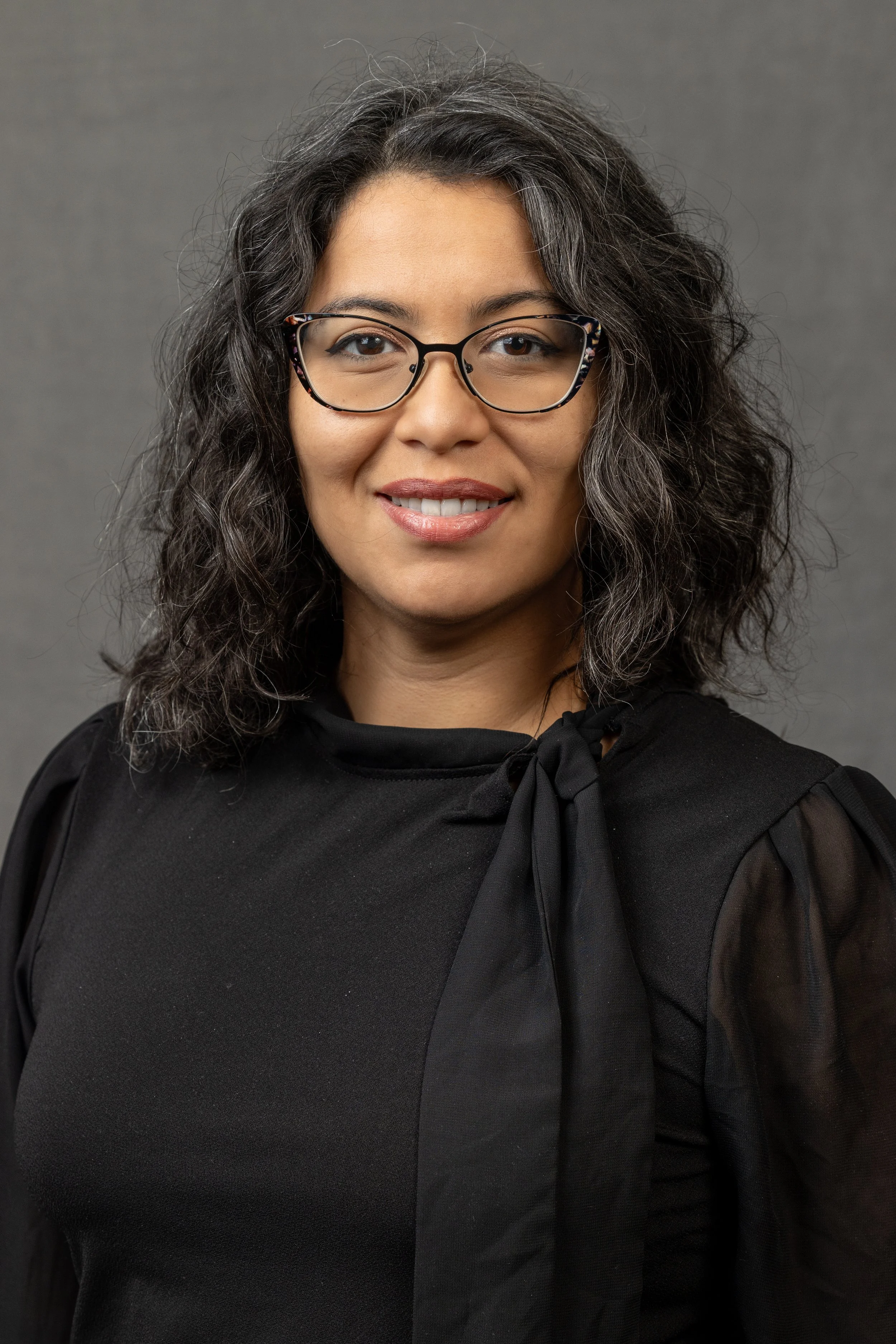 A woman with curly dark hair and glasses smiling in front of a gray background.
