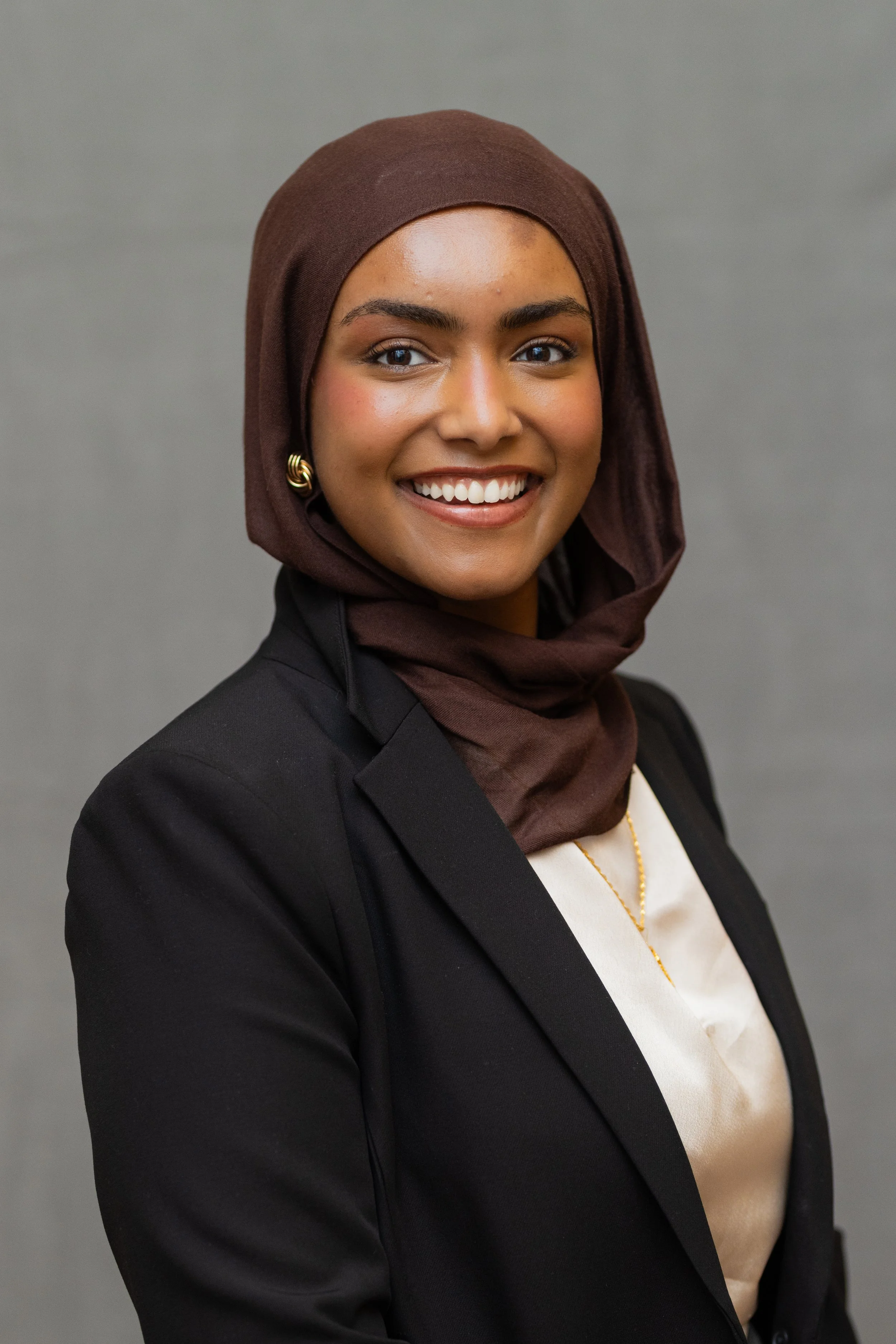 A young woman with medium brown skin, wearing a dark brown hijab, black blazer, white blouse, gold necklace, and earrings, smiling in front of a gray background.