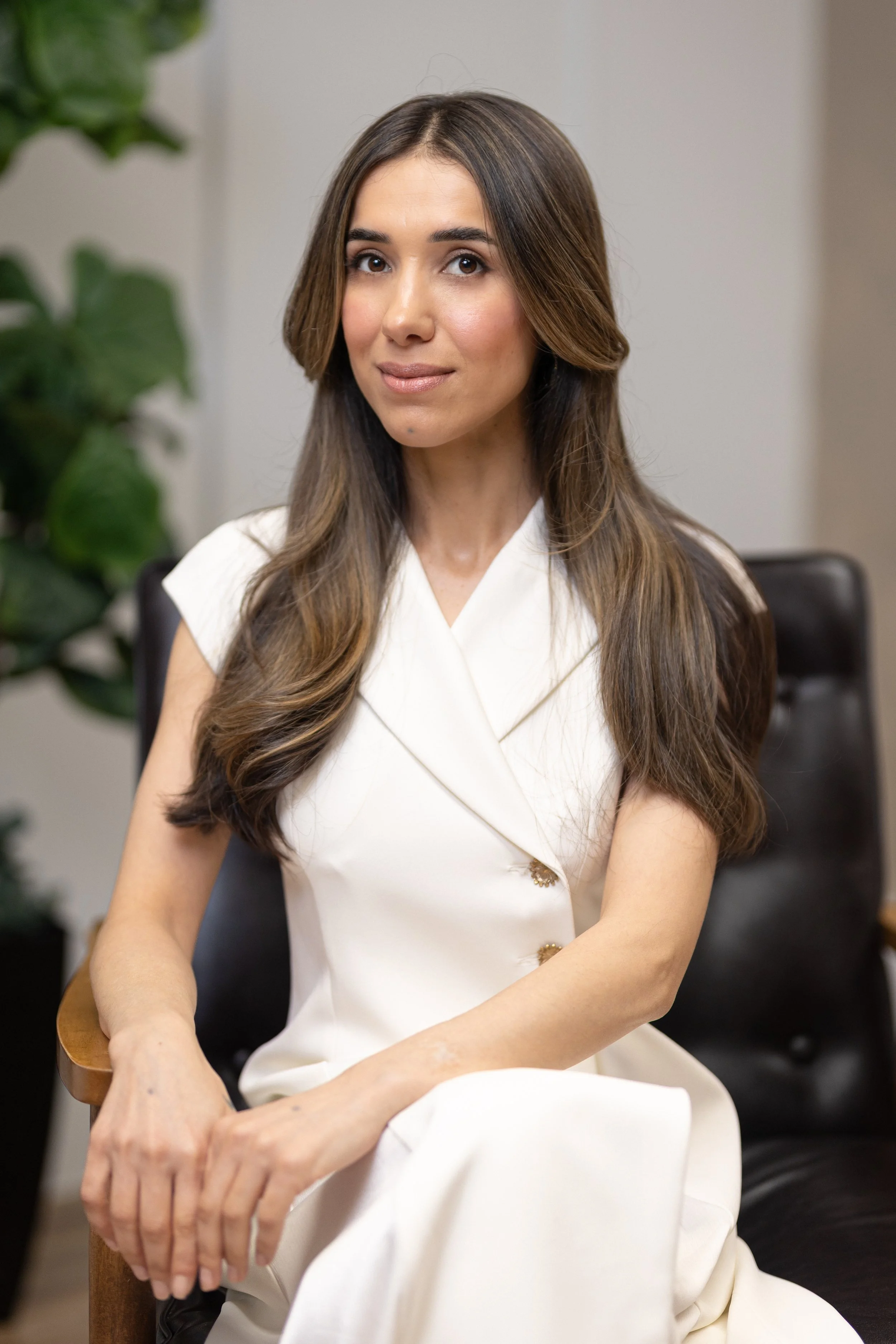 A woman with long brown hair and light skin sitting on a black leather chair, wearing a white dress, in an indoor setting with a plant in the background.