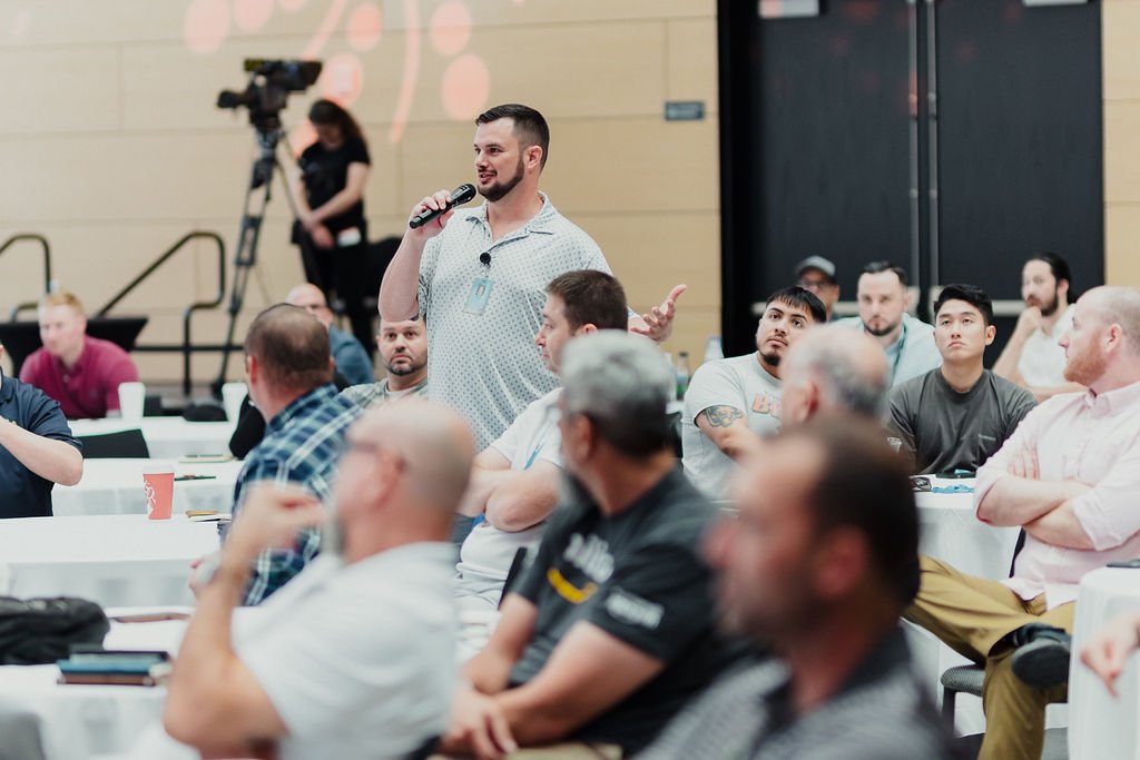 A man is speaking into a microphone during a conference with an audience seated at tables, including diverse individuals, some taking notes, in a large indoor hall.
