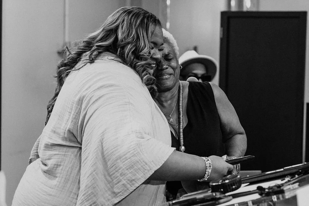 Two women embrace and look at a phone together, appearing happy and emotional.