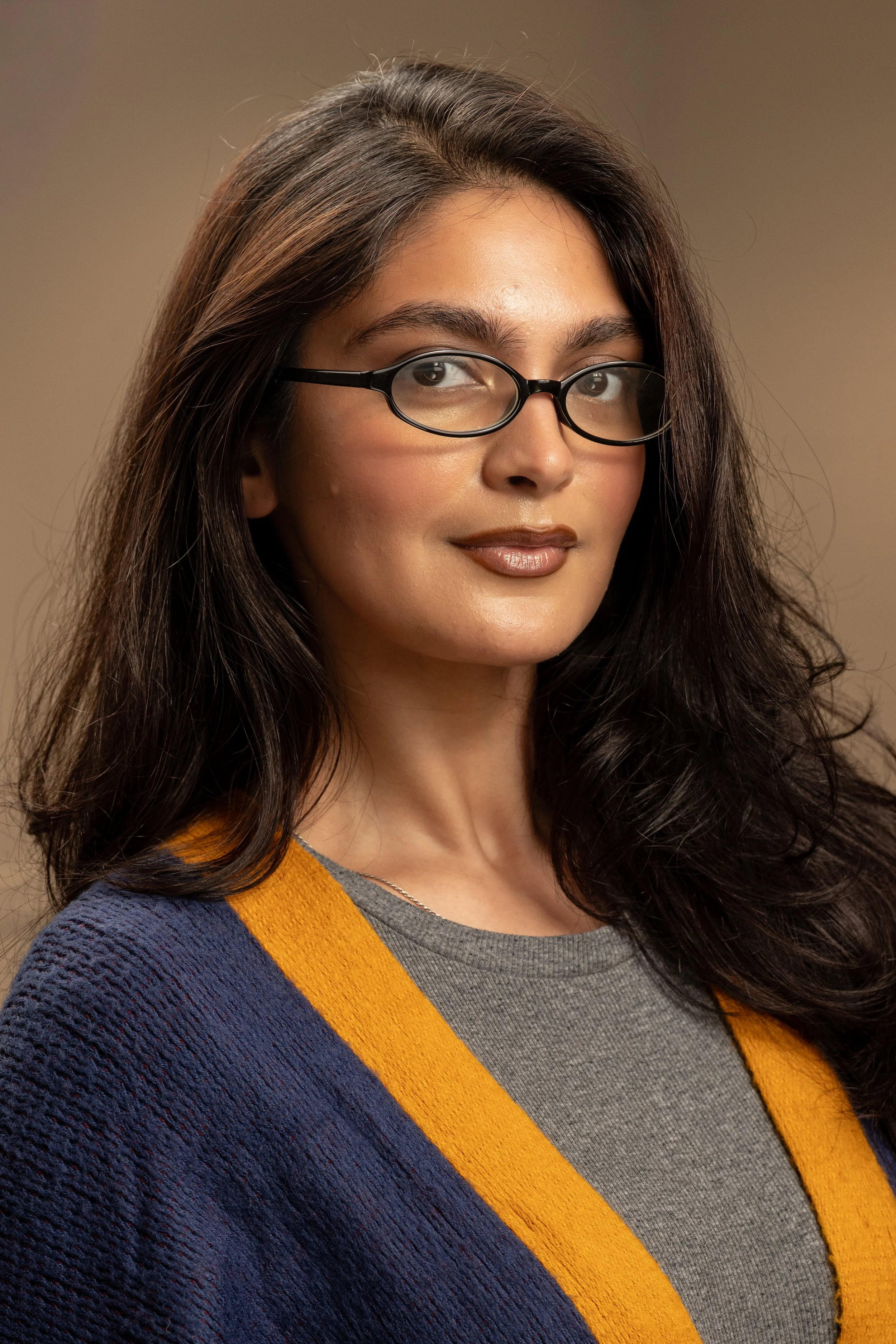 A woman with long dark hair wearing glasses, a gray shirt, and a blue cardigan with yellow trim, posing against a neutral background.