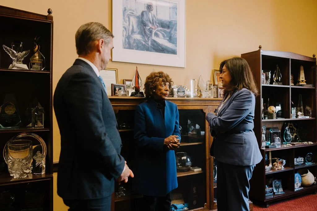 Three people engaged in conversation inside an office, with glass display cabinets and framed pictures on the walls.