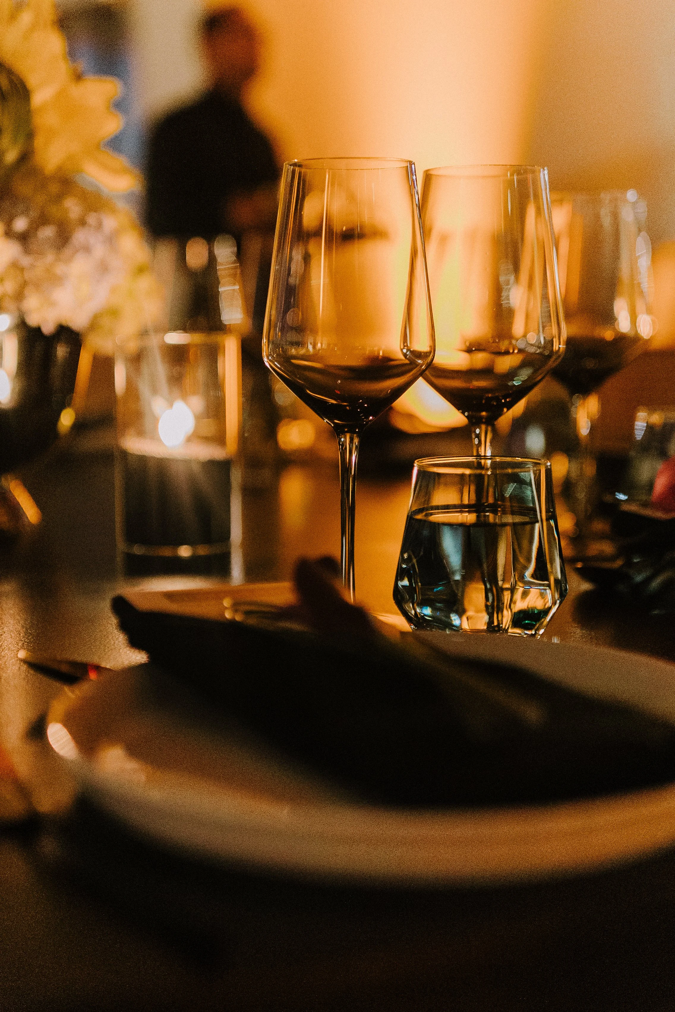 Close-up of wine glasses and a glass of water on a restaurant table, with blurred background of a person and dim lighting.