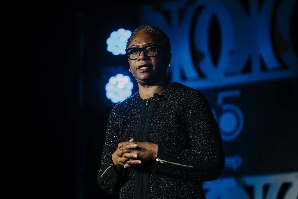 A woman with short hair, glasses, and earrings speaking on stage at a conference or event, with a dark background and blue stage lights.