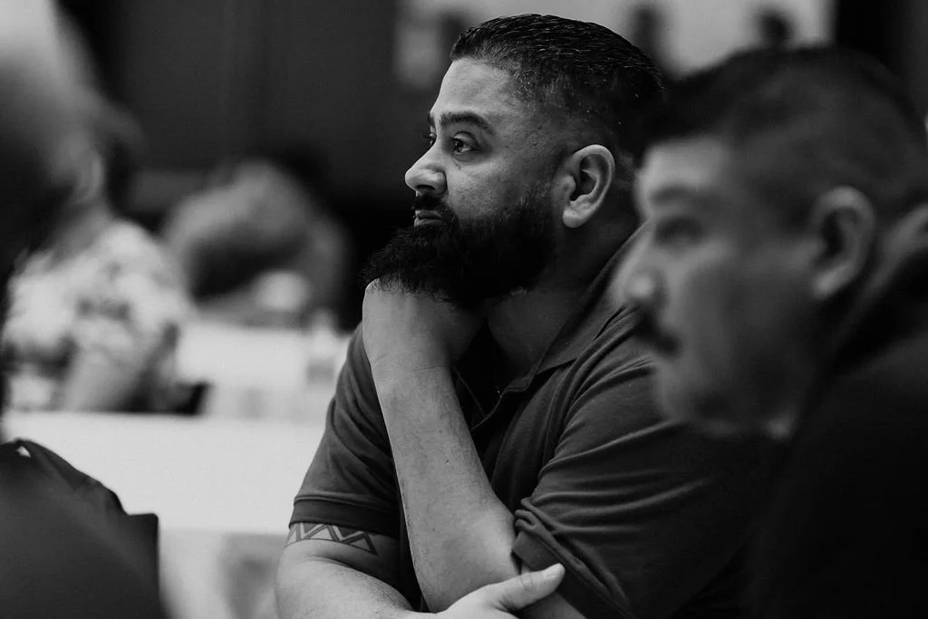 A man with a beard and neatly styled hair, sitting at a table, resting his chin on his hand, in a black and white photo during a meeting or conference.