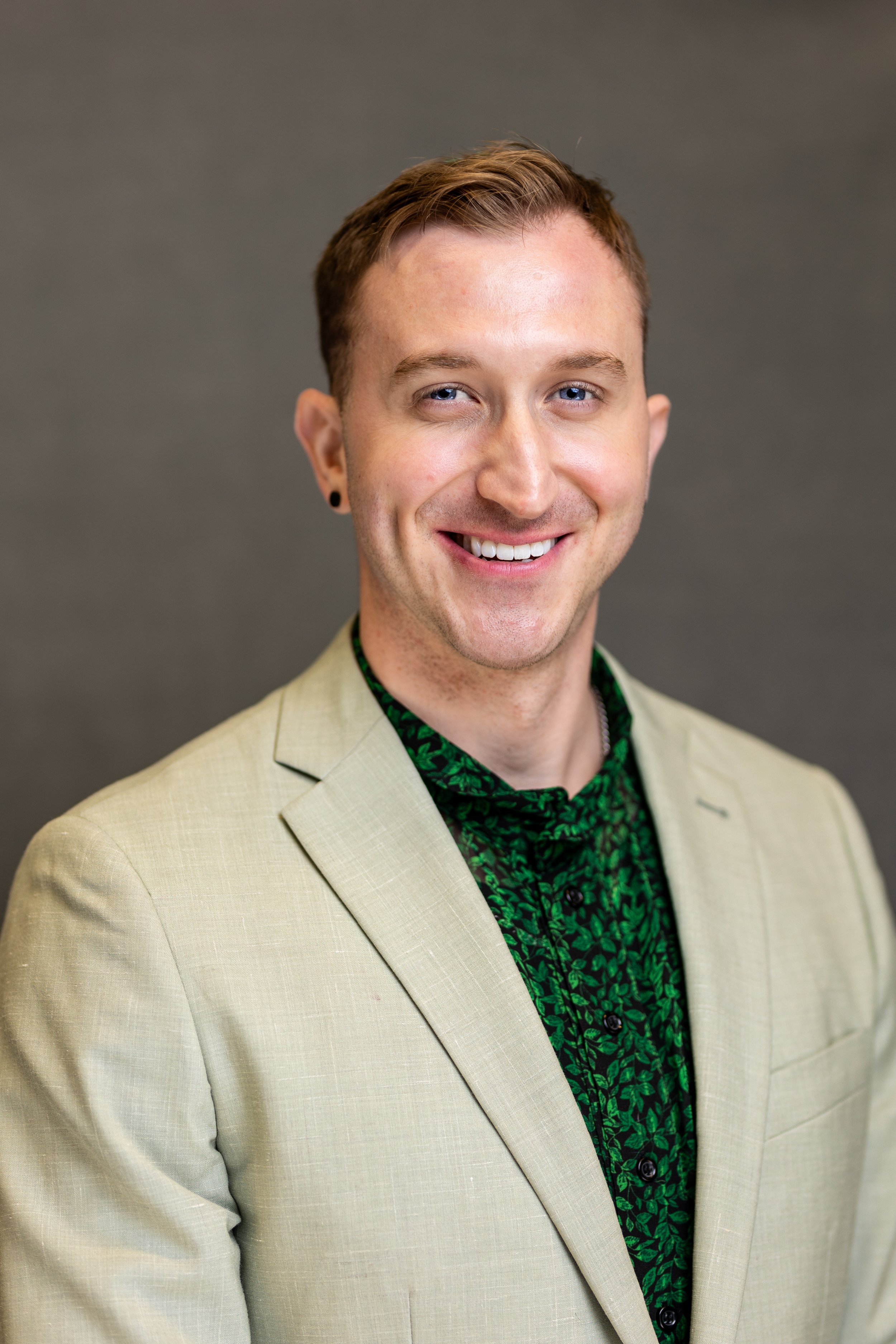 A portrait of a smiling man with short brown hair, light skin, wearing a beige blazer and a dark green patterned shirt, standing against a gray background.