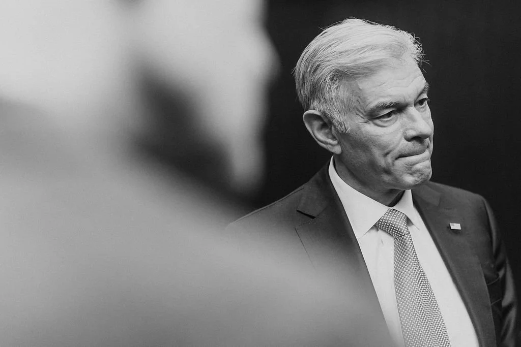 A black and white photo of a man with gray hair in a suit and tie, looking thoughtful. The background is dark, and part of another person's shoulder is visible in the foreground.