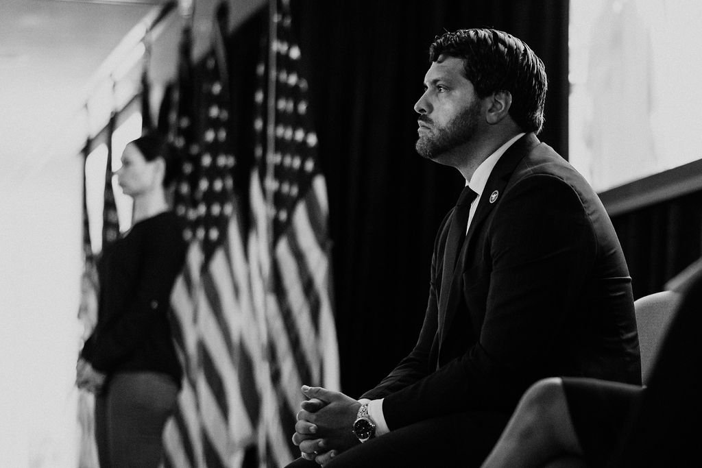 A black and white photo of a man in a suit sitting and looking serious. In the background, a woman is standing near several American flags.