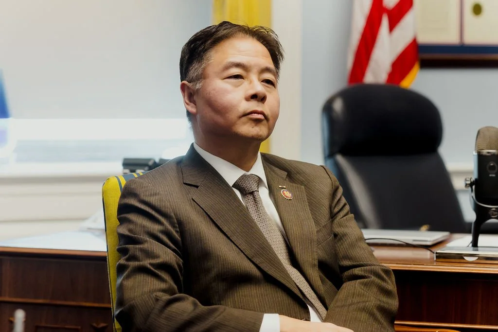 A man in a brown suit sitting in a chair in a conference room, with an American flag in the background.