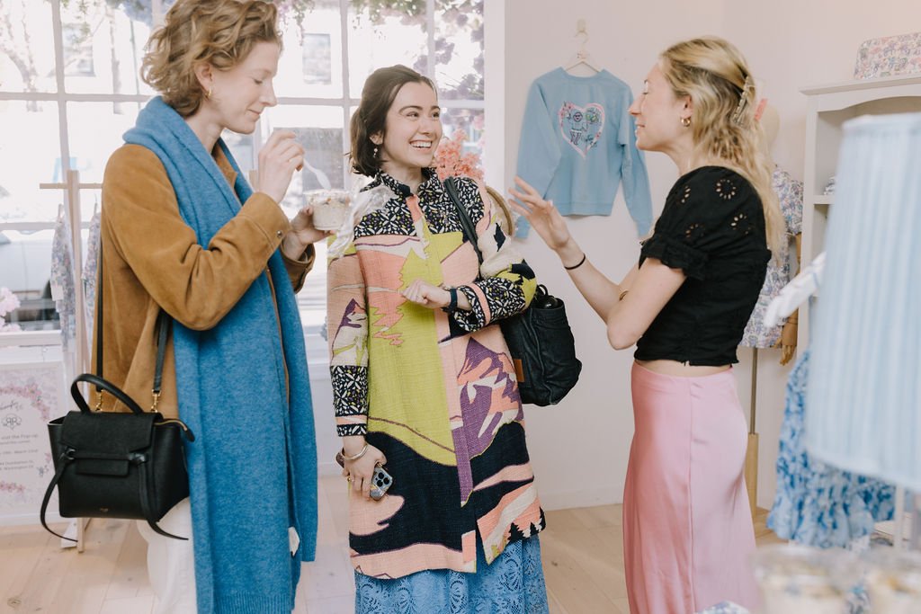 Three women engaging in conversation inside a boutique or craft shop, with one woman smiling and gesturing, and the other two listening, with clothing and crafts visible in the background.
