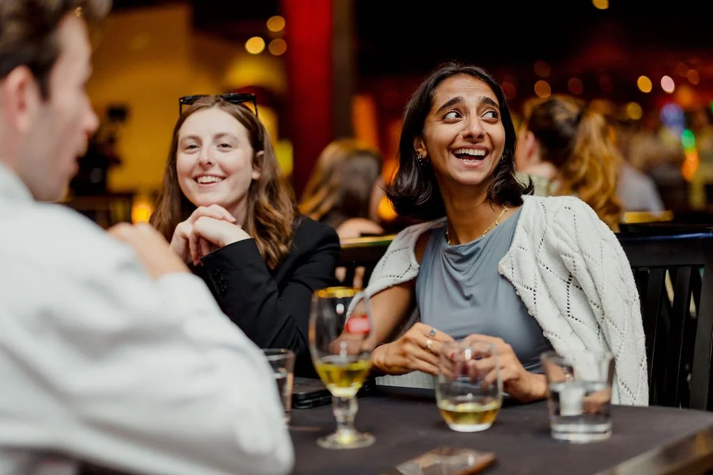 Three people sitting at a table in a restaurant, smiling and engaging in conversation. Two women are visible, one with short dark hair wearing a light gray top and white cardigan, and the other with long brown hair and black blazer. A man is partiall