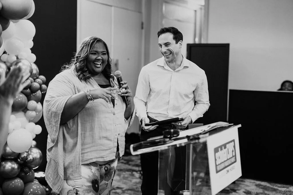 A woman with curly hair holding a microphone smiling, standing next to a man in a white shirt laughing while holding a tablet at an event with balloons and a podium.