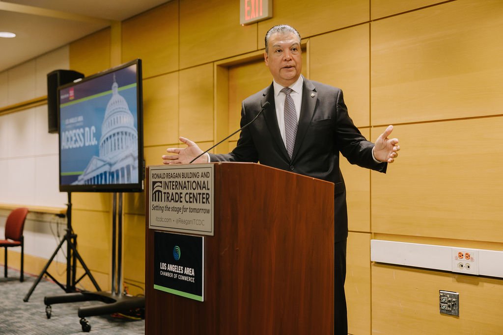 A man in a suit speaking at a podium during a conference at the Ronald Reagan Building and International Trade Center. There is a large screen displaying a Capitol building image and the text 'ACCESS DC' beside him.