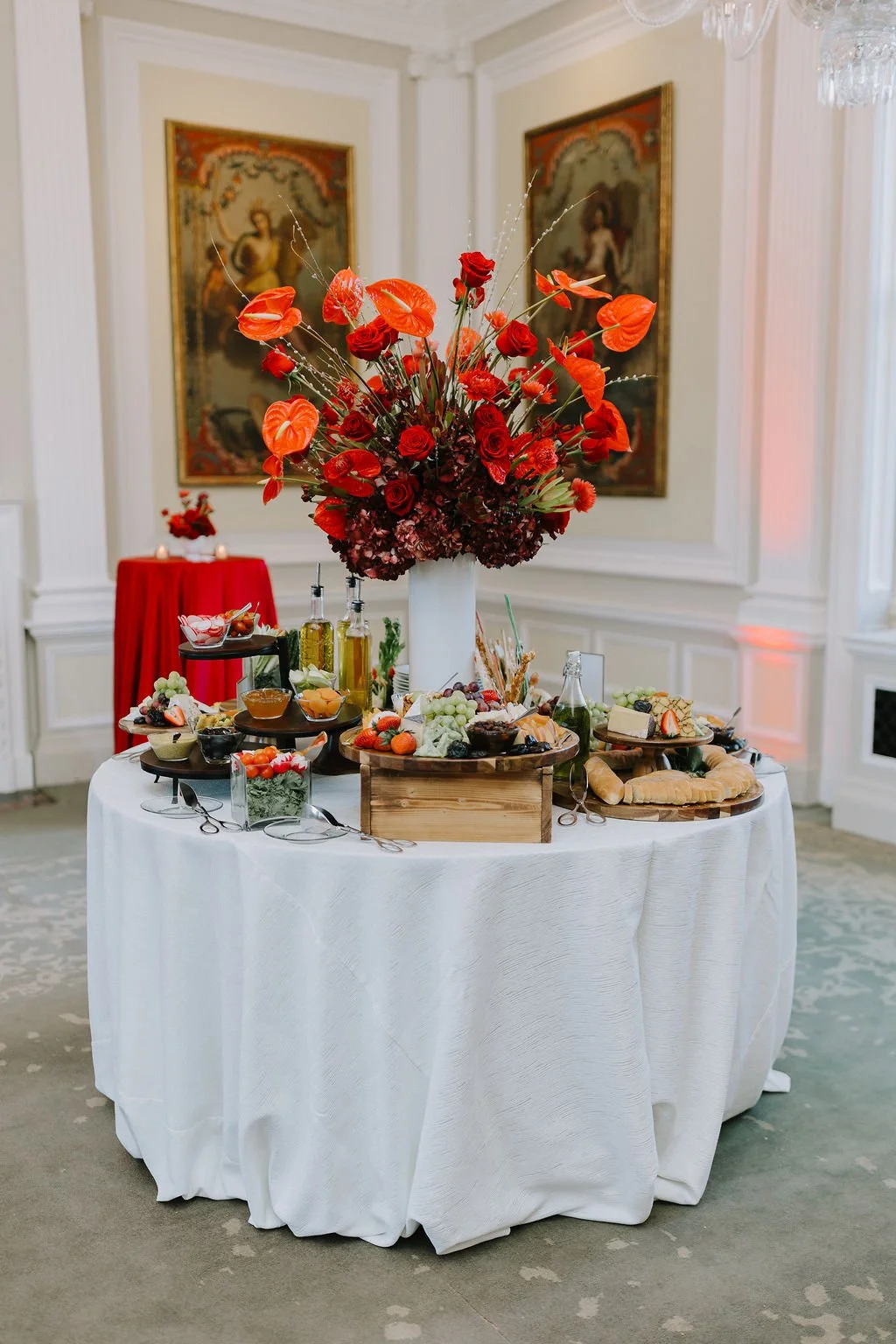 A table with a white tablecloth displays a large floral arrangement of red and orange flowers, surrounded by plates of fruits, cheese, bread, and beverages. In the background, there are paintings on the wall and a small red table with a red flower ar