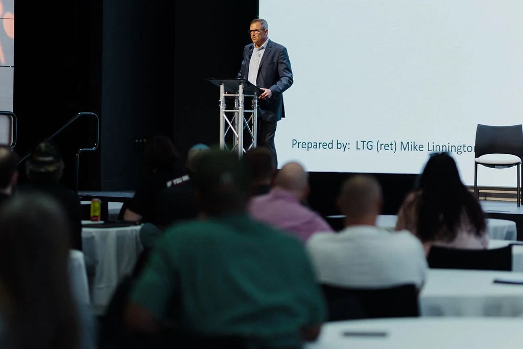 Man in a suit speaking at a podium during a presentation in a conference room.
