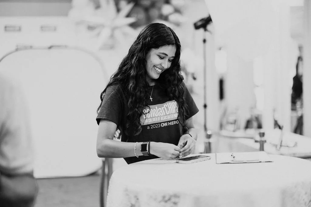 A woman with long dark curly hair smiling and looking down at a table, wearing a dark t-shirt and a watch, with a cross necklace, in a bright indoor setting.