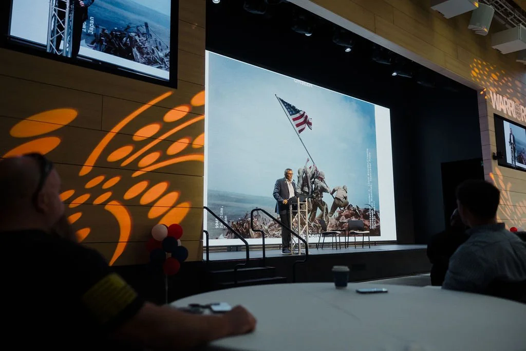 A speaker standing at a podium with a large image of soldiers raising the American flag on a hill behind him during a presentation or conference.