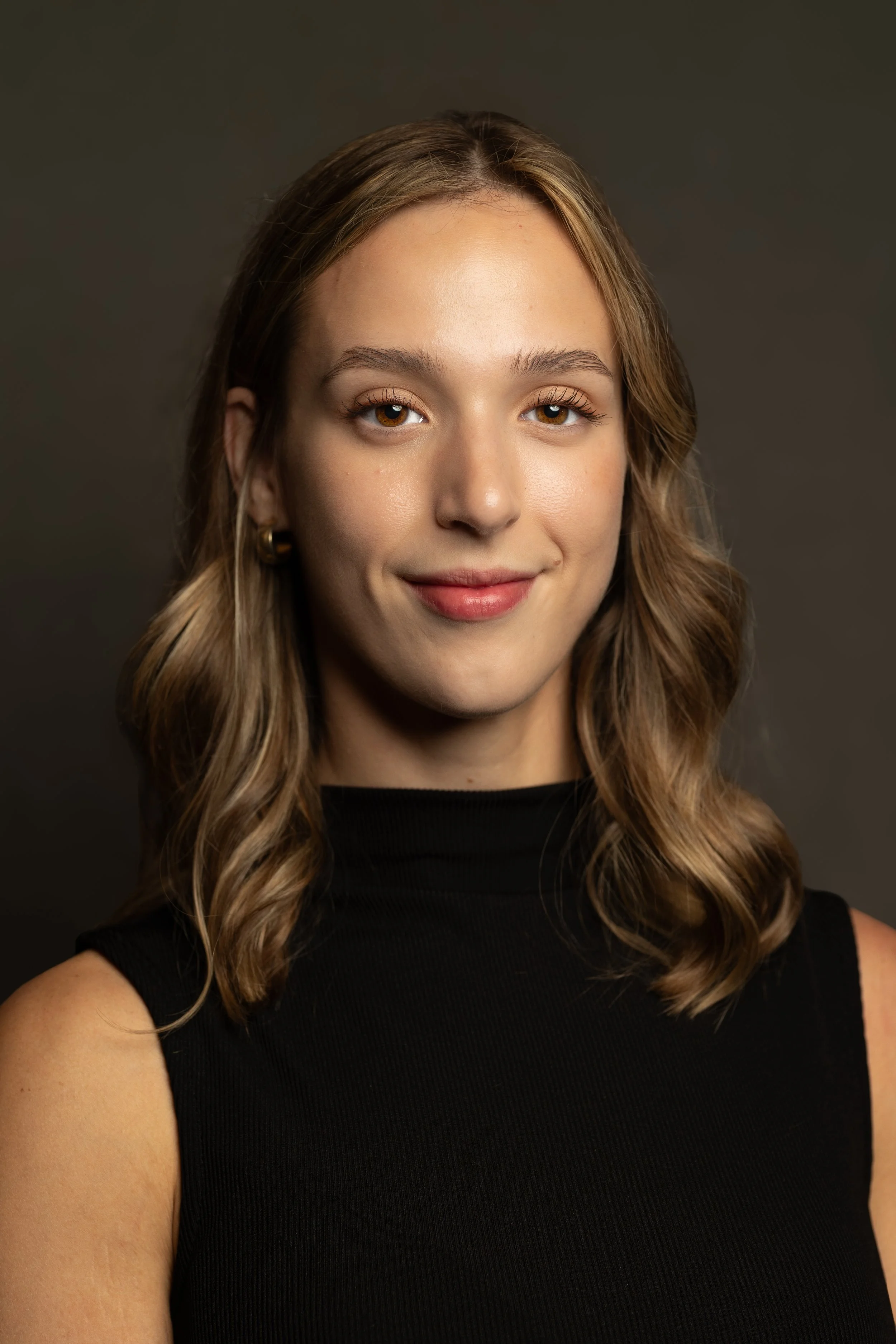Portrait of a young woman with wavy brown hair, wearing a black sleeveless top and small gold hoop earrings, smiling gently against a dark background.