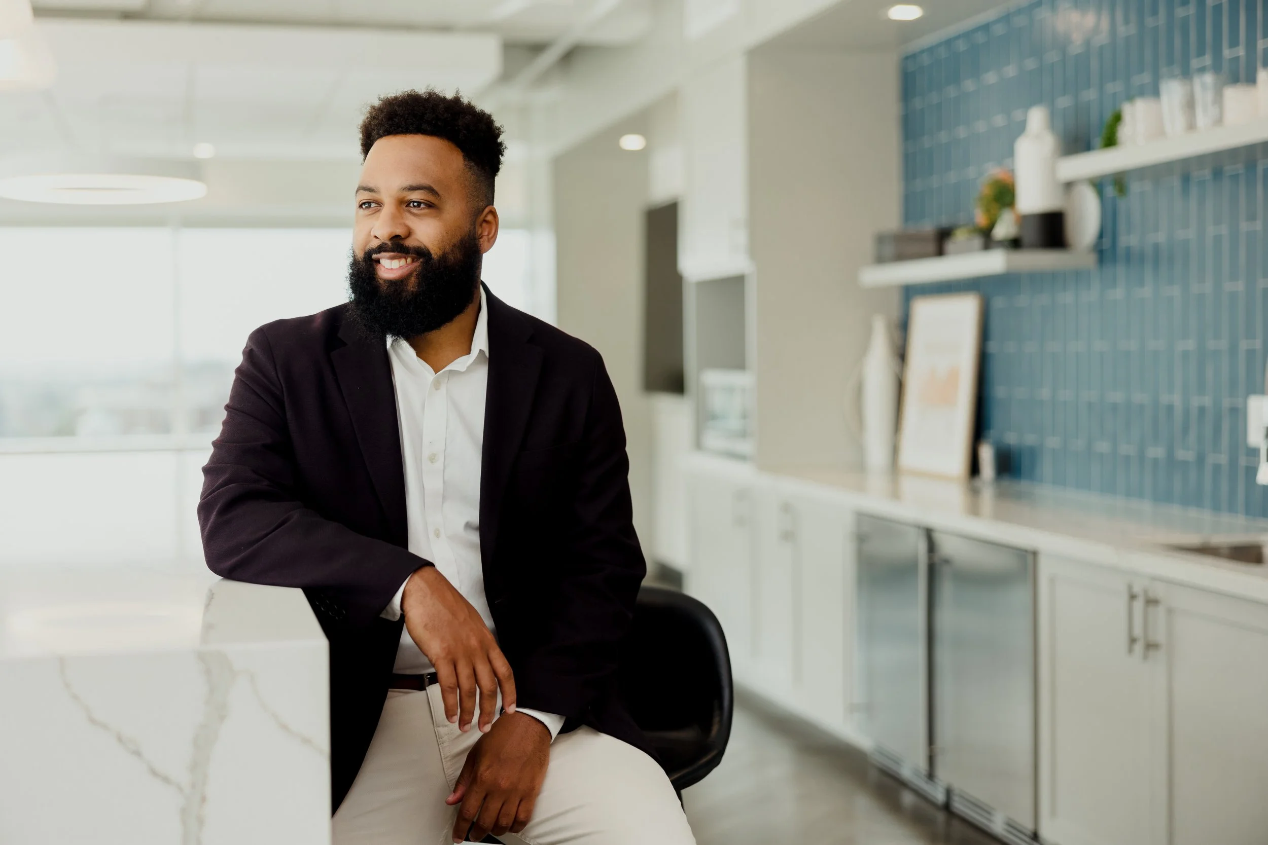 A man with a beard wearing a black blazer and white shirt sitting in a modern office kitchen space.