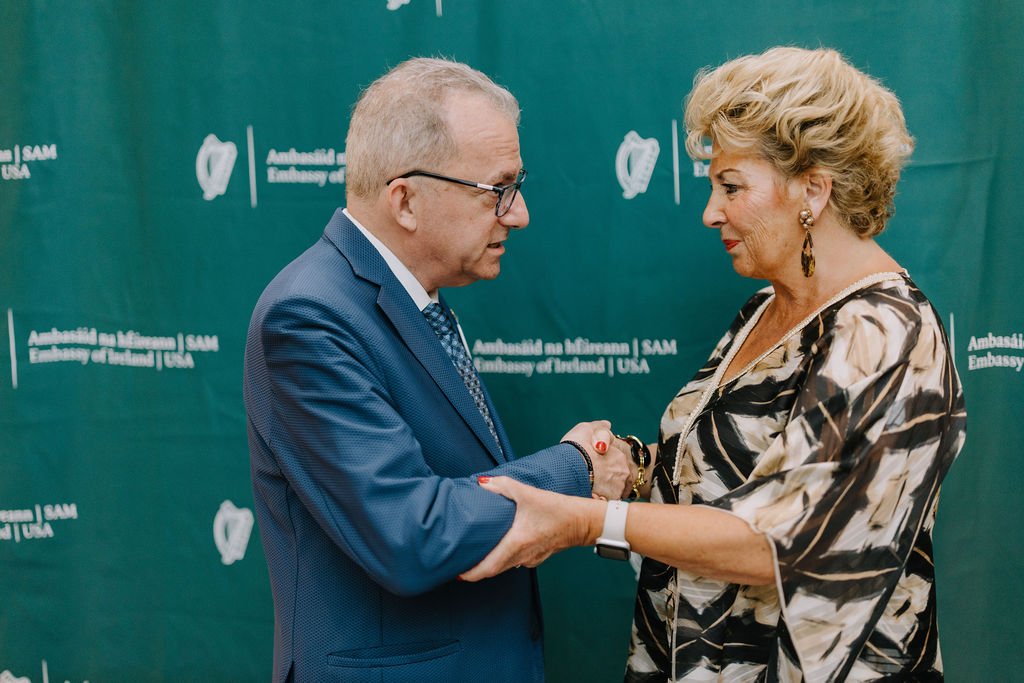 A man and a woman shaking hands at an event with a green backdrop that has logos and text for the Embassy of Ireland and SAM USA.