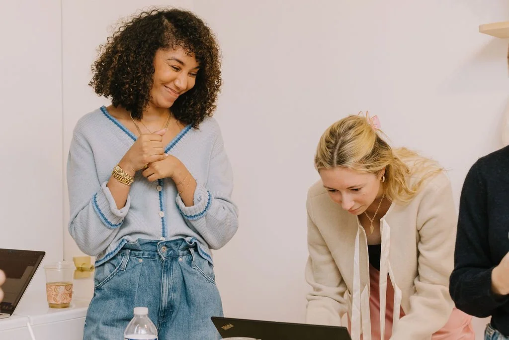 Two women in a casual office environment, one smiling and the other focused on a laptop.