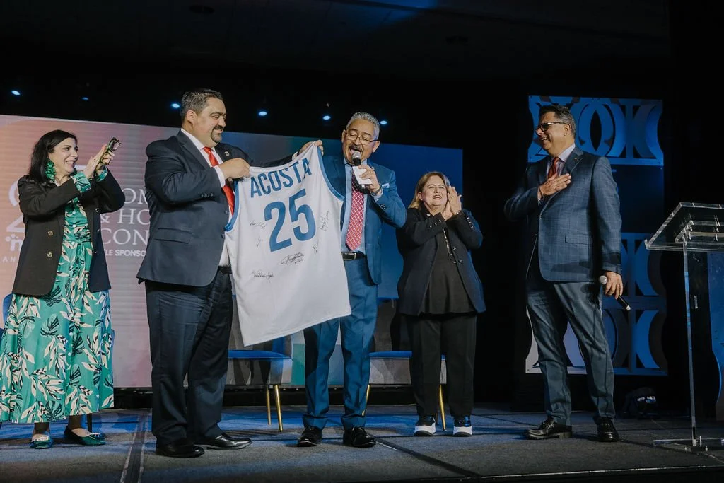 Group of five people on stage celebrating a sports jersey, with one person holding a microphone, at an indoor event.