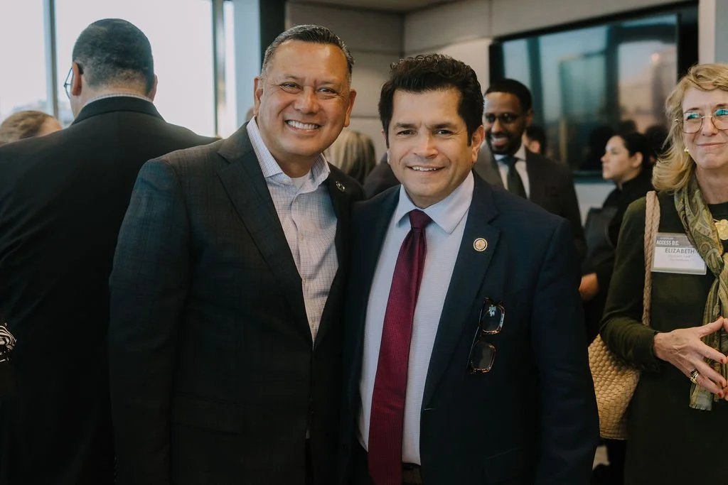 Two men in suits smiling at a professional event, standing close together, with a woman and other attendees in the background.