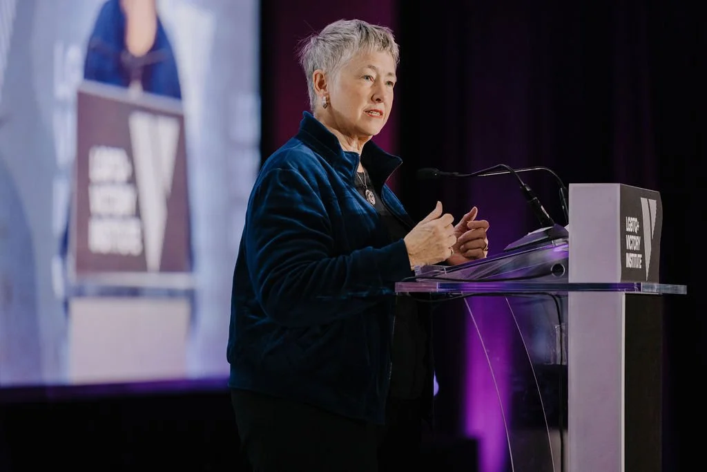 An older woman with short gray hair is speaking at a podium during a conference or event, with a large screen behind her displaying a blurred image.