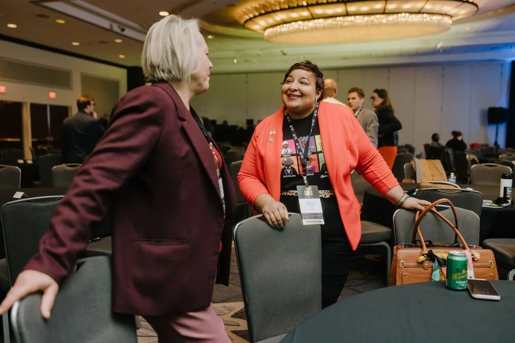 Two women engaged in conversation at a conference or event, with chairs and other attendees visible in the background.