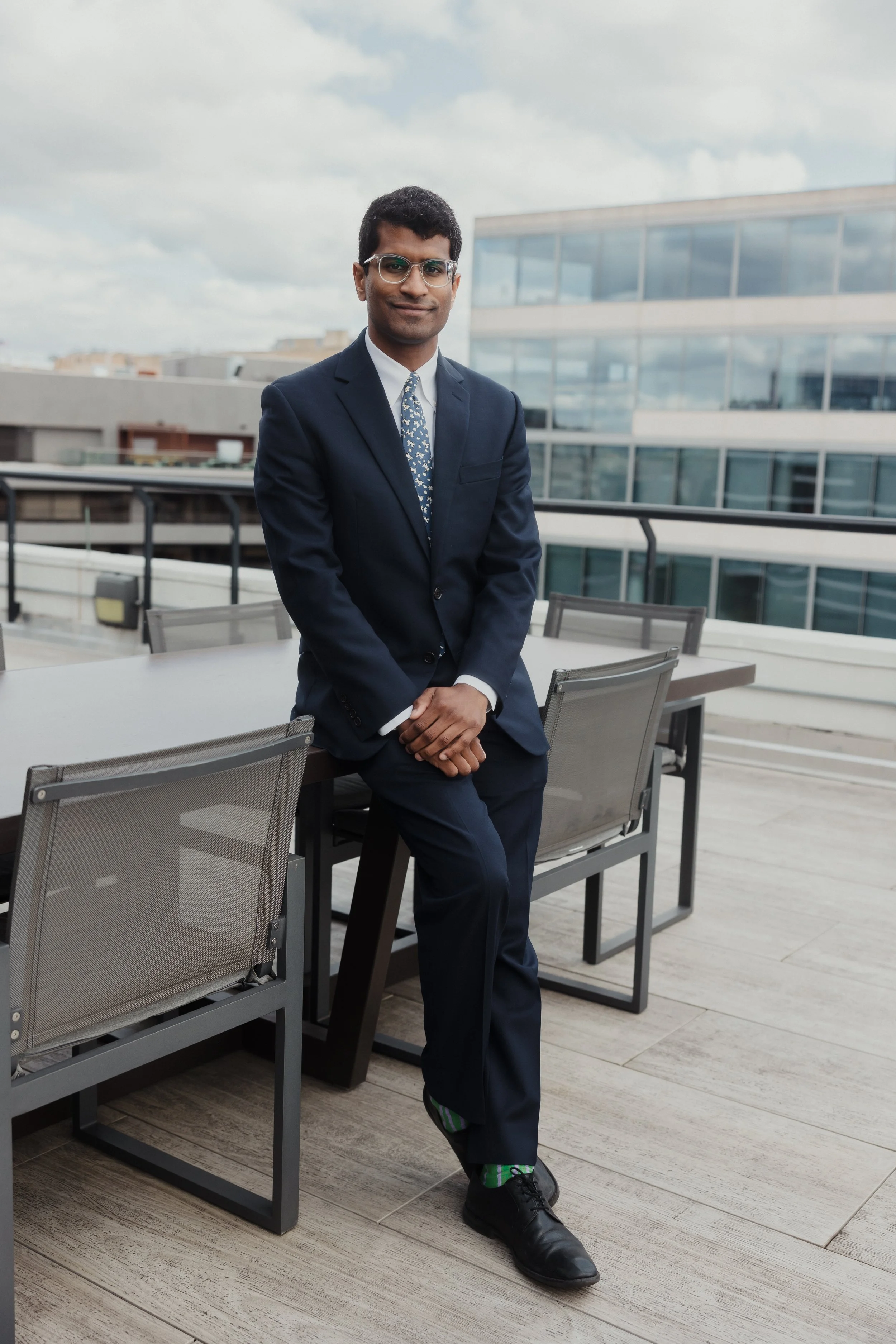 A man in a navy suit and patterned tie with glasses poses on a balcony with modern buildings in the background.