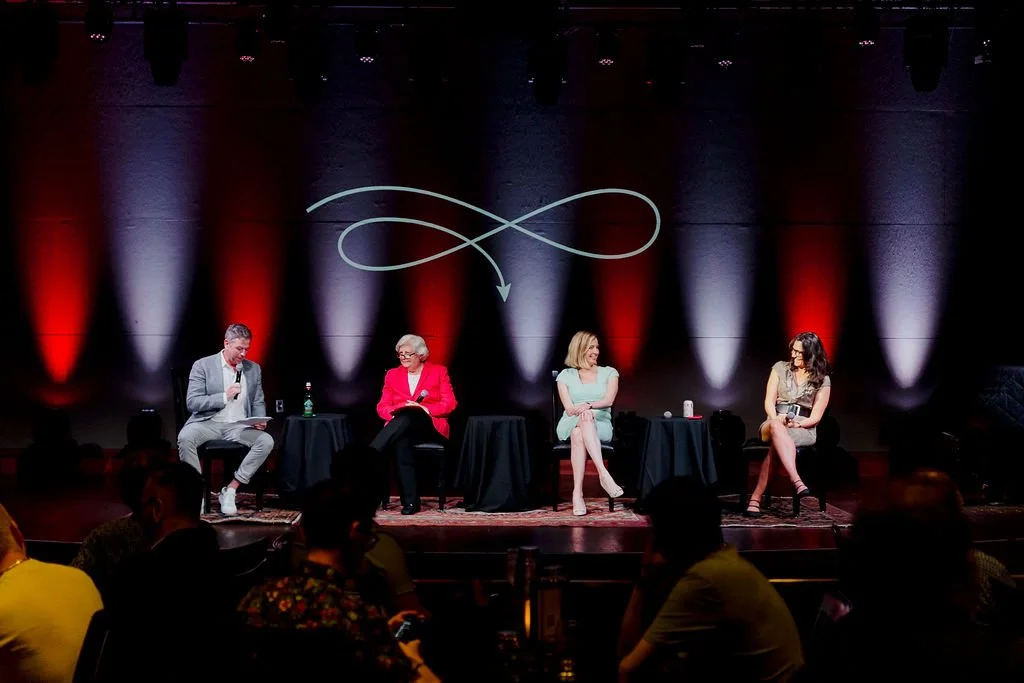 Four people seated on stage during a panel discussion, with a dark background illuminated by red and purple lighting, and a decorative line projection.