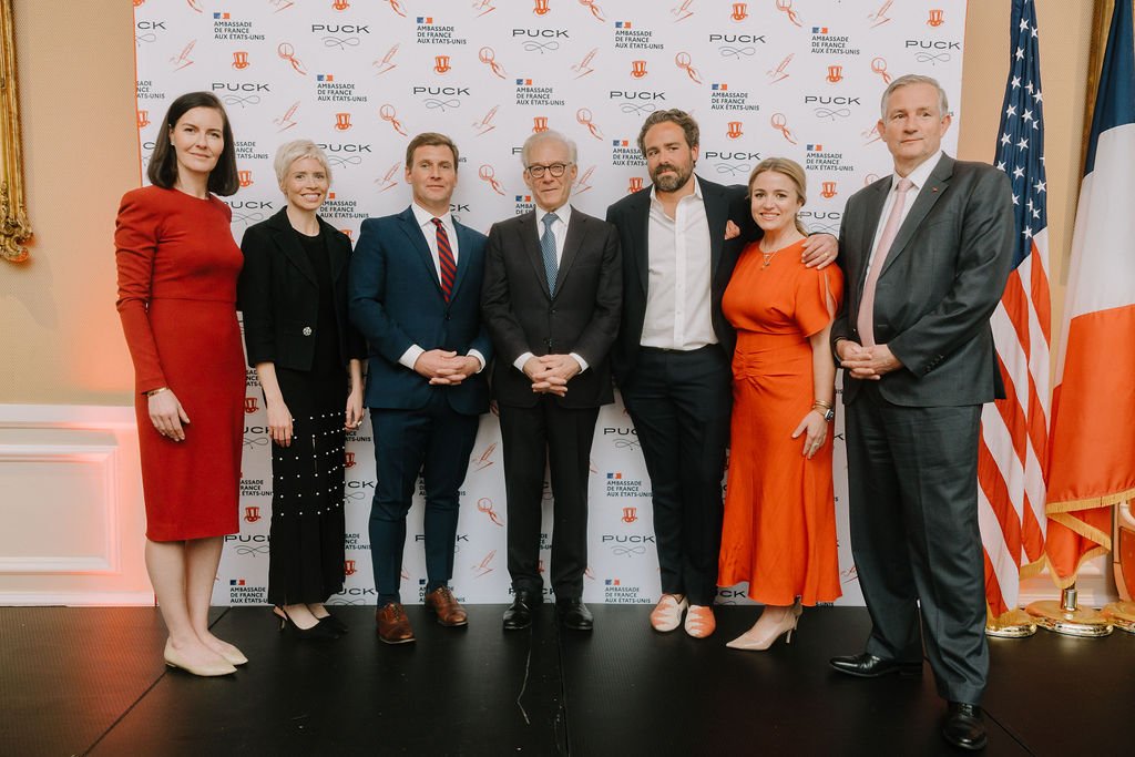 Group of seven people dressed in formal attire posing in front of a backdrop with logos and flags, including the United States and France flag, at a formal event.