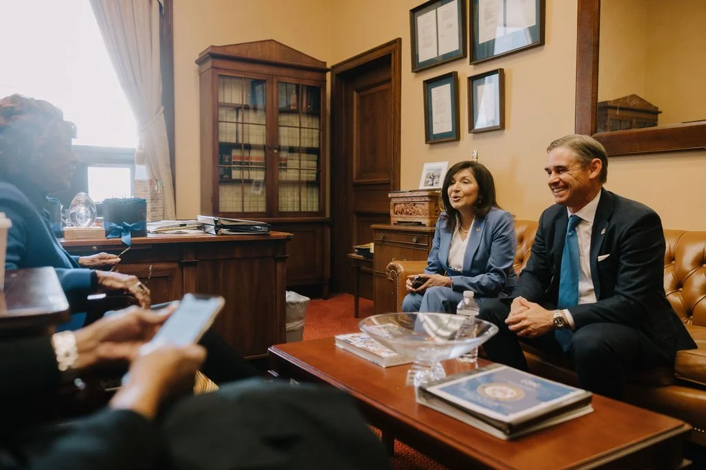 A professional meeting in an office with five people. Two are sitting on brown leather sofas, smiling and engaging in conversation. One woman in a blue blazer is holding a phone, and a man in a dark suit with a blue tie is sitting next to her. Three 