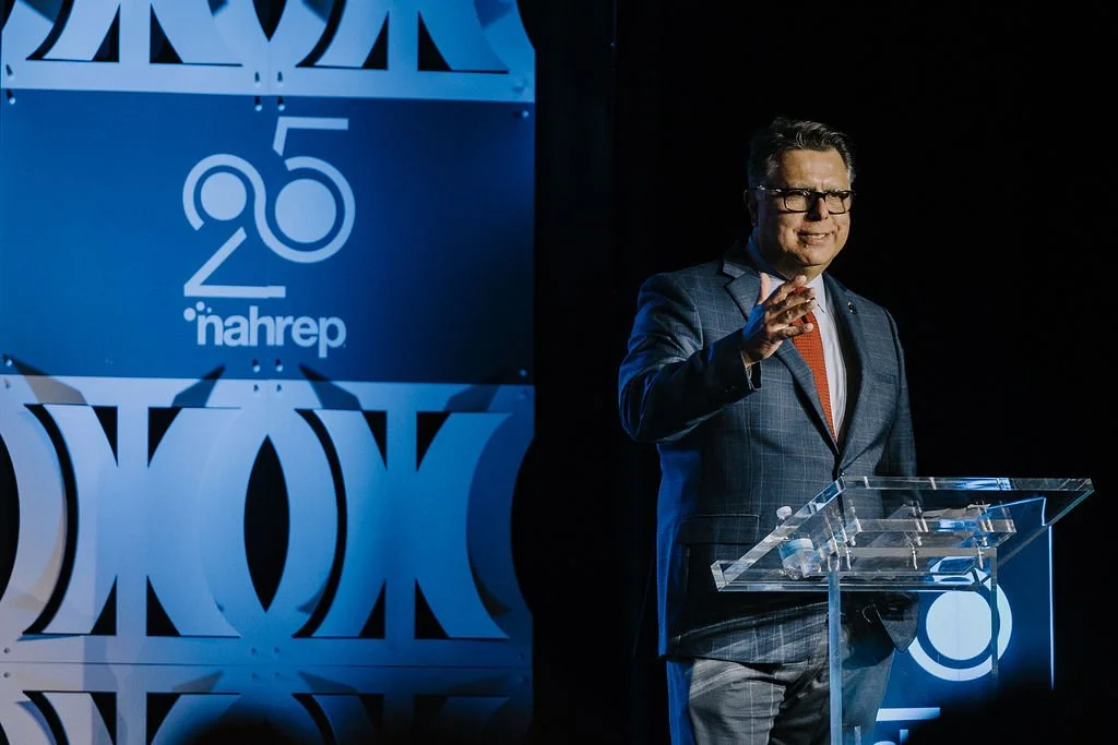 A man in a gray suit and red tie standing behind a clear podium, speaking at an event with a large blue and white backdrop that features the number 95, the word 'nahrep,' and a stylized logo.