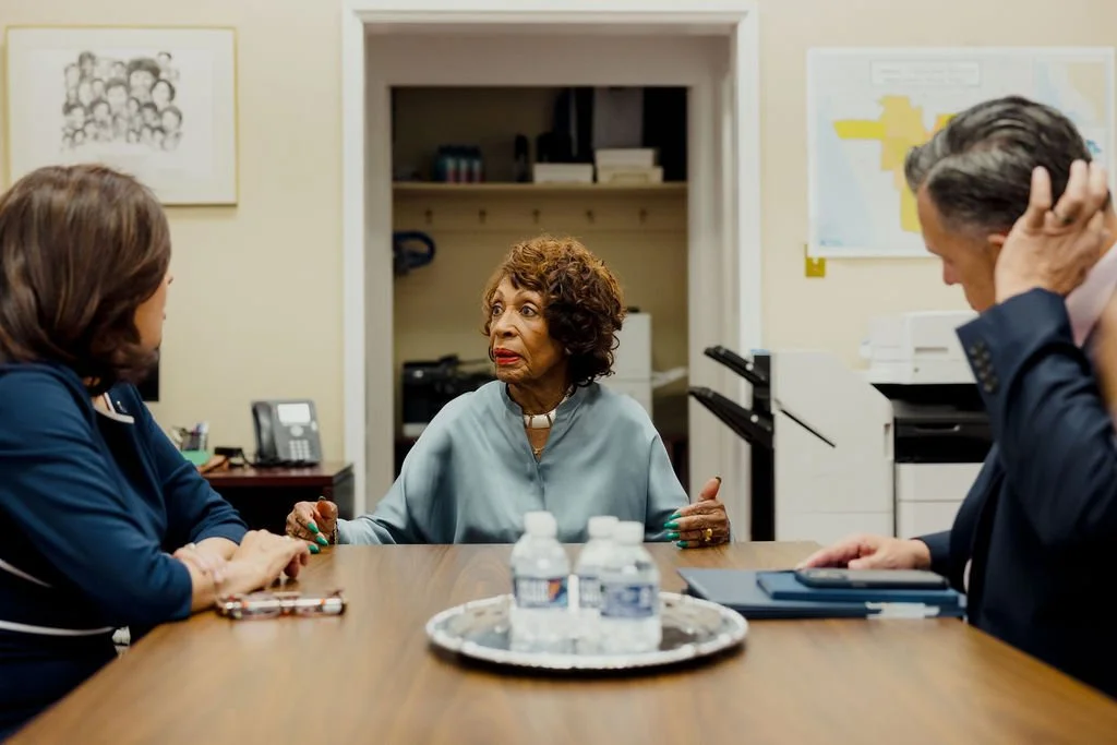 Three people in a meeting room having a discussion. A woman in the middle appears to be speaking and gesturing, while a woman on the left and a man on the right listen attentively.
