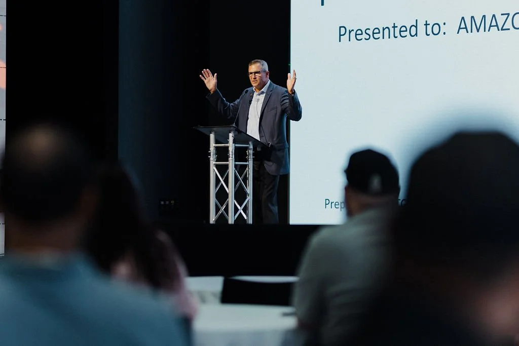 A man in a suit giving a presentation on a stage with a microphone stand, addressing an audience.