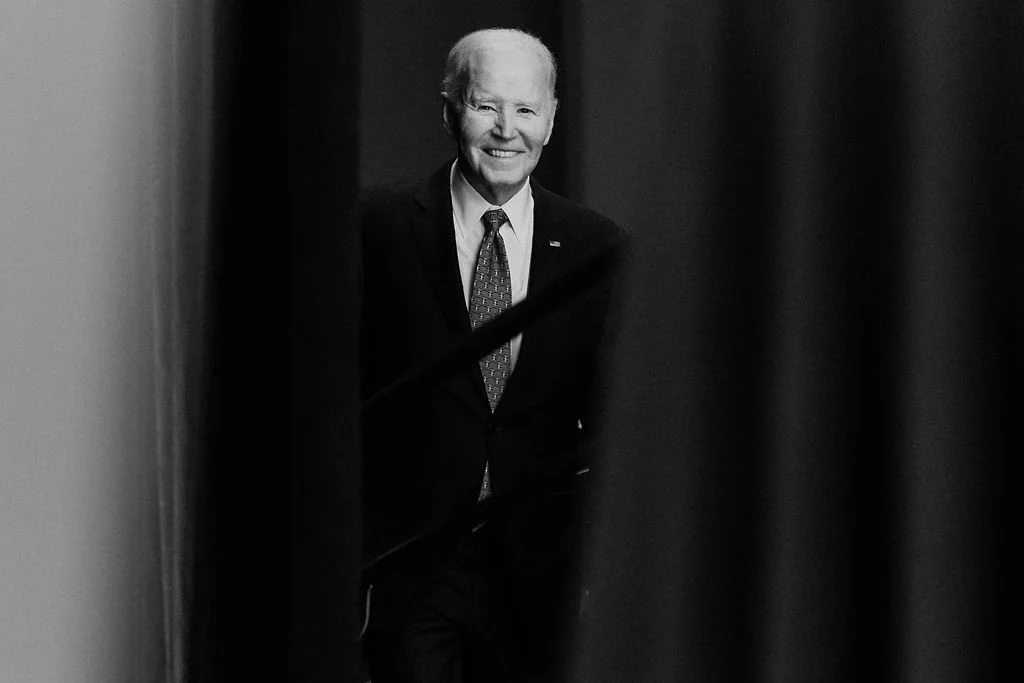 Black and white photo of Joe Biden smiling, wearing a suit and tie, peeking through curtains.