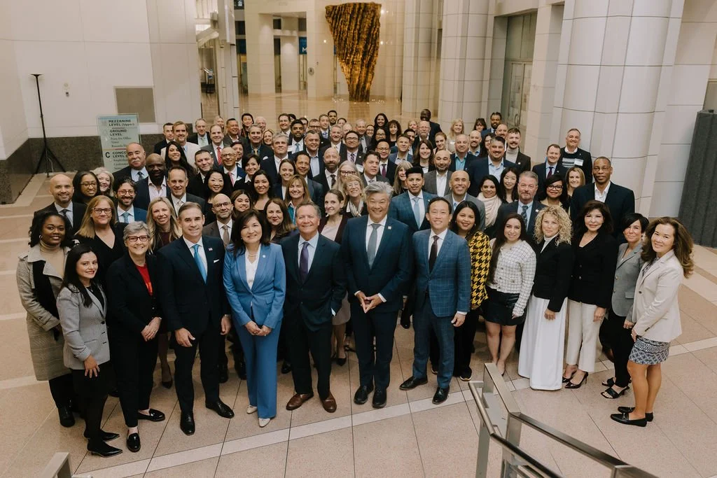 A large group of diverse business people in formal attire gathered in a spacious, modern office lobby or atrium for a group photo.