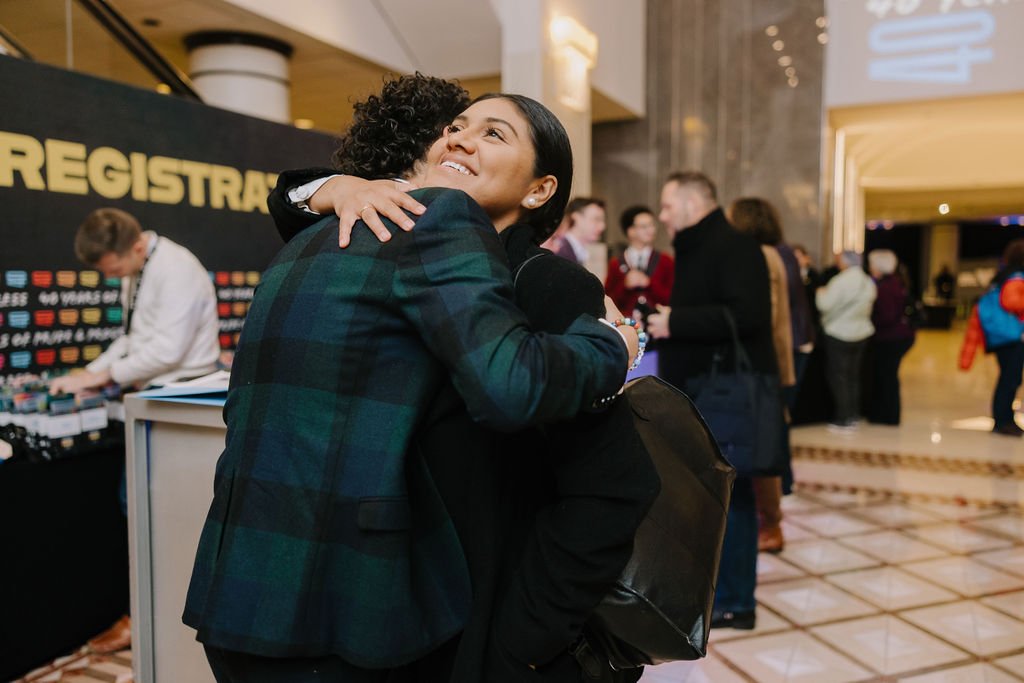 Two women hugging at a busy conference or event, with people and a registration desk in the background.