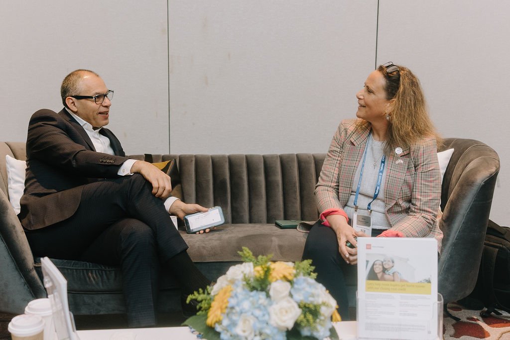 A man and woman sitting on a gray sofa, engaged in conversation, with a flower arrangement on the table in front of them.