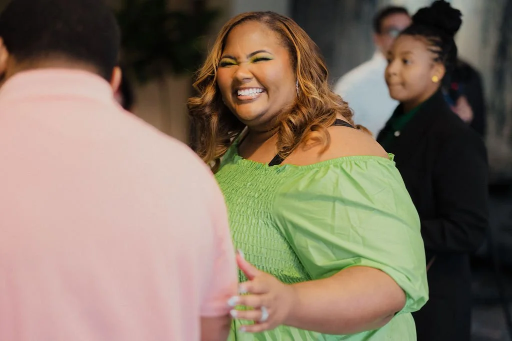 A woman with long, wavy hair smiling joyfully at a social gathering, wearing a green off-shoulder top.