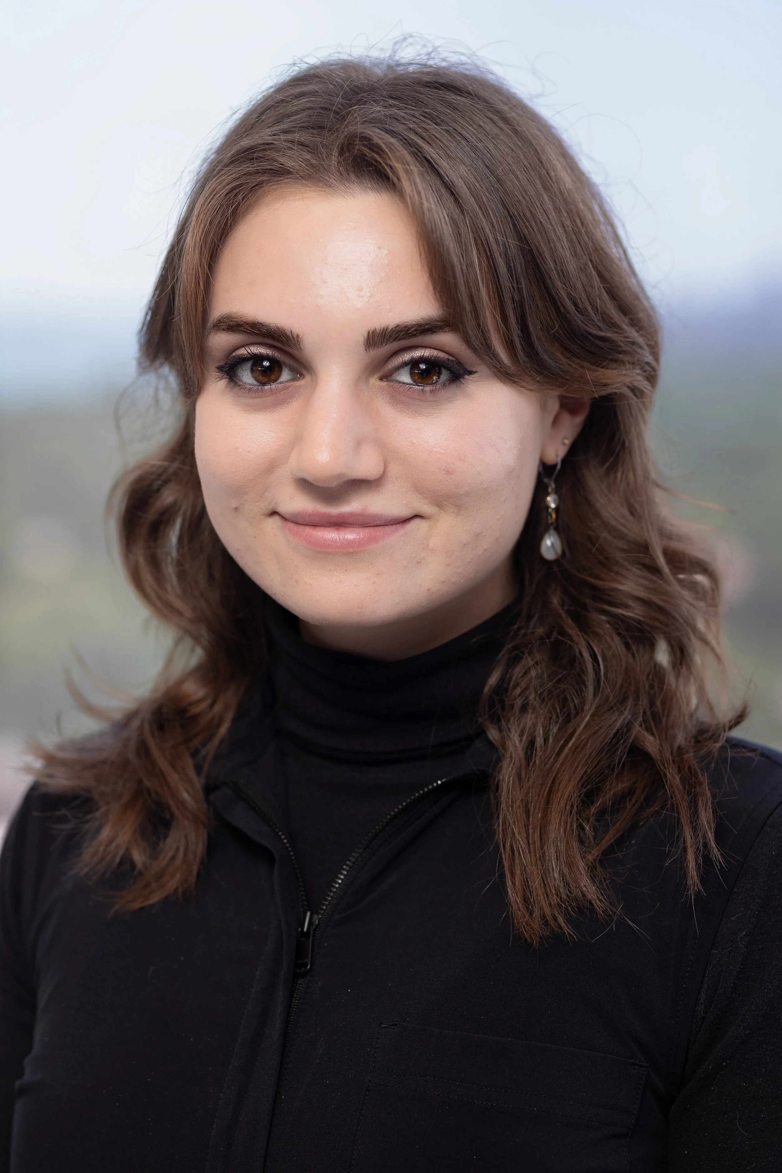A woman with shoulder-length brown hair, wearing a black turtleneck and earrings, smiling at the camera with a blurred outdoor background.