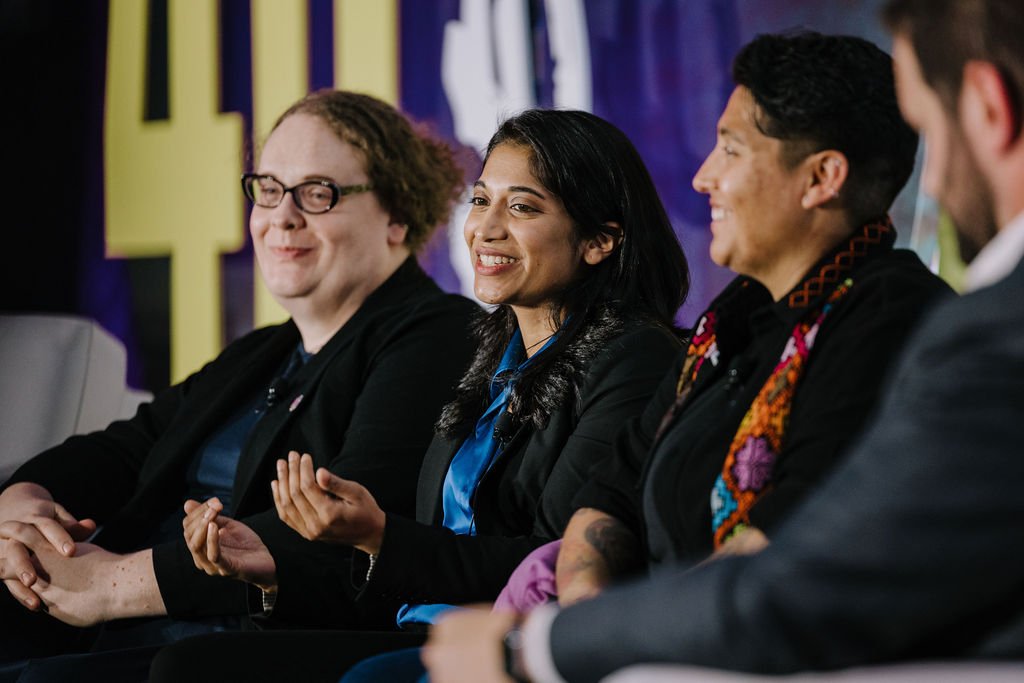 Four diverse individuals sitting on a panel, smiling and engaging in a discussion at an event.