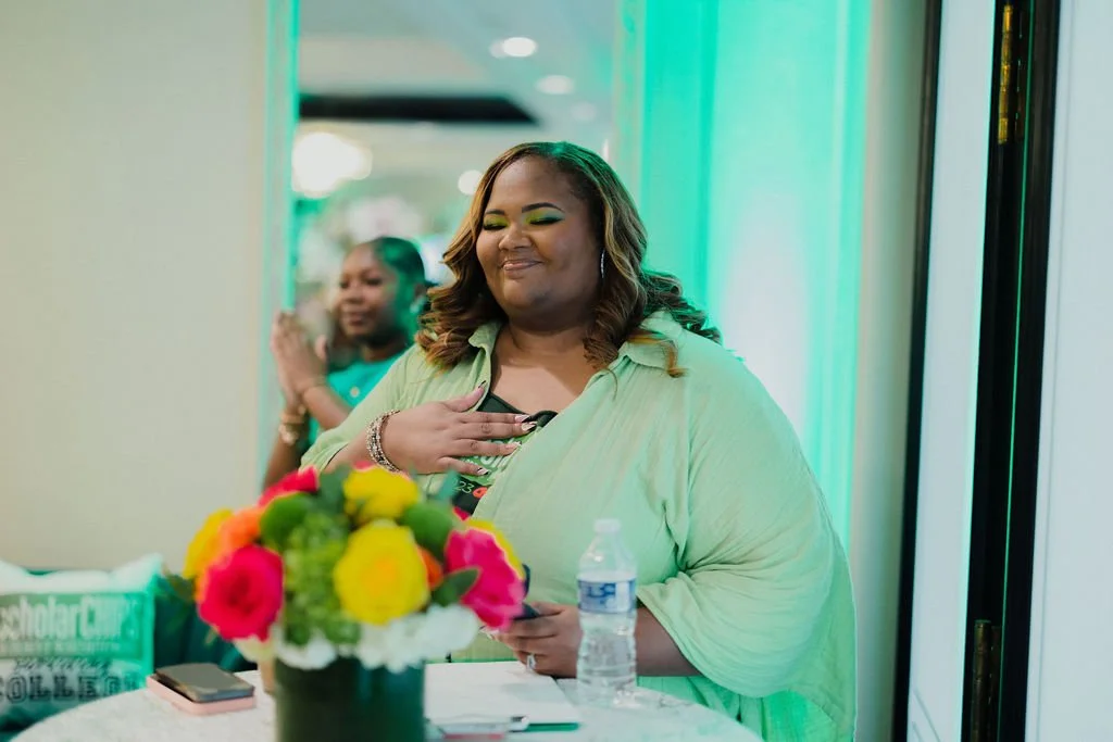 A woman standing in front of a table with a colorful flower arrangement, holding a phone, with a water bottle on the table, and another woman in the background clapping, in a room with green lighting.