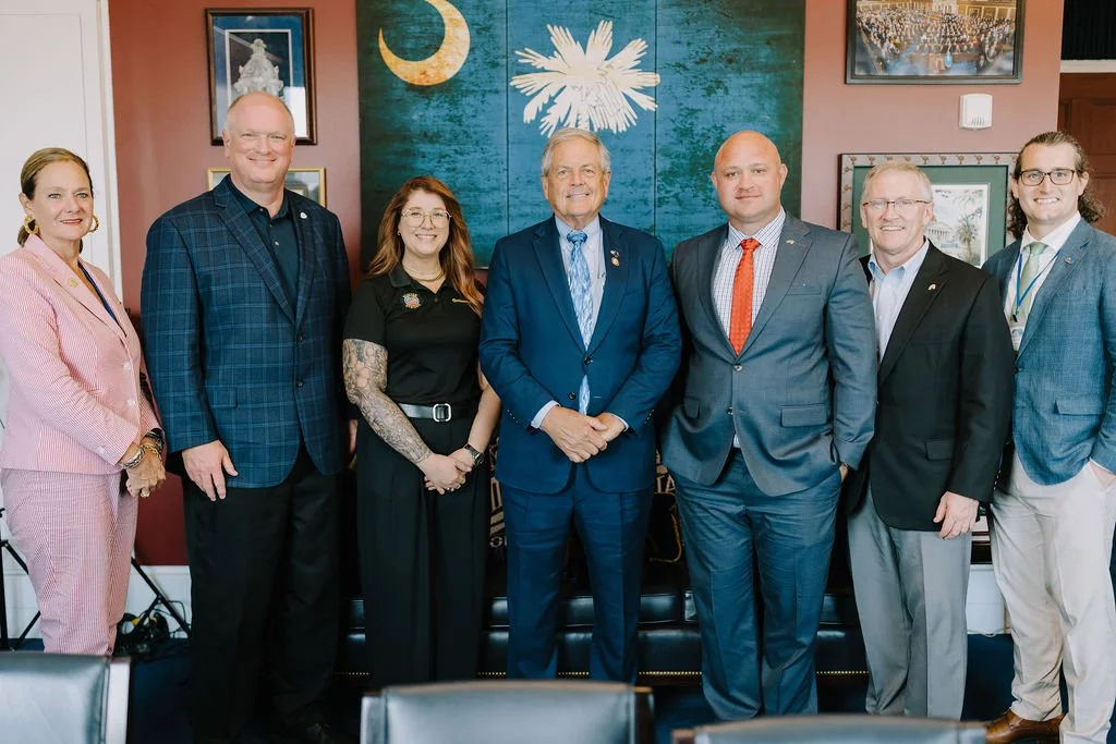 A group of eight professionally dressed people standing side by side in a room with artwork and a blue background with a white eagle emblem and a crescent moon. They are smiling and posing for the photo.