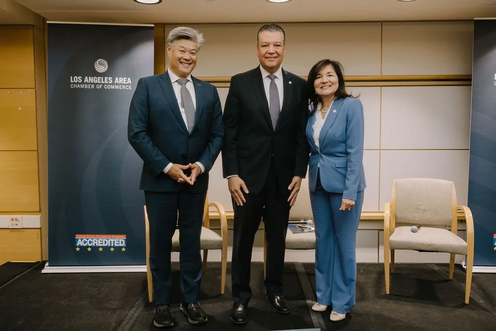 Three professionally dressed individuals standing together in front of Los Angeles Area Chamber of Commerce banners, smiling at the camera.