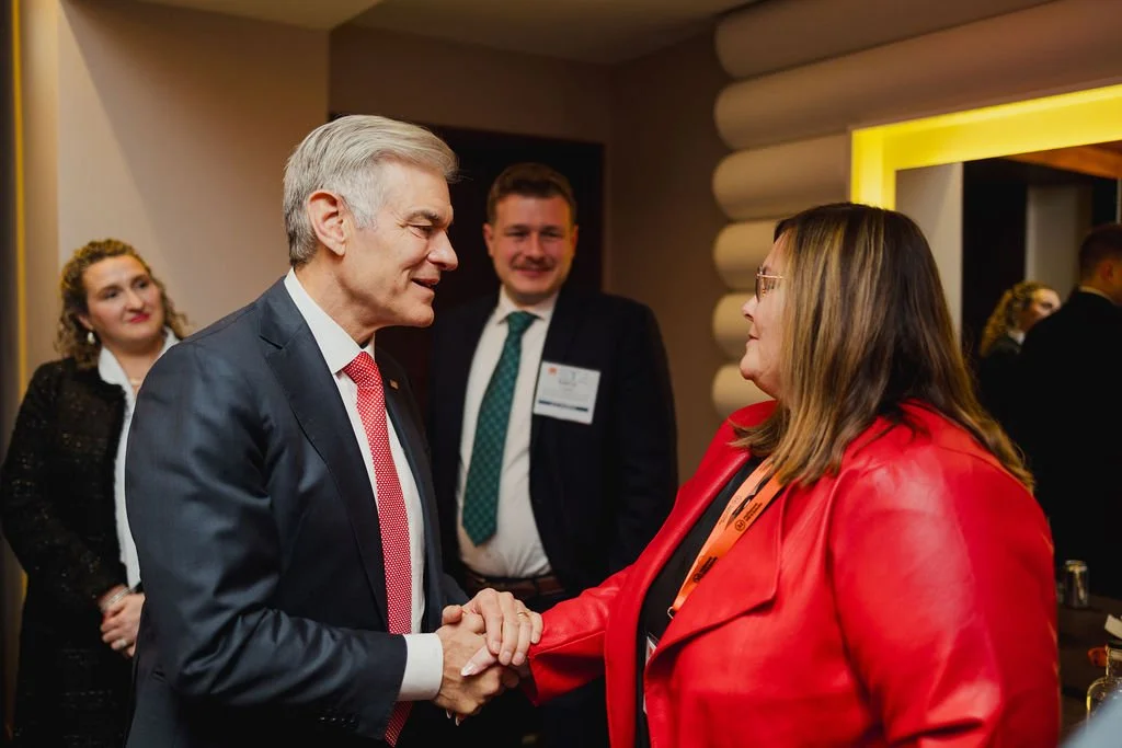 A man and a woman shaking hands at a professional event. The man has gray hair and is wearing a dark suit with a red tie. The woman has brown hair, glasses, and is wearing a red jacket. There are other people in the background, dressed professionally, in a warmly lit conference or reception area.